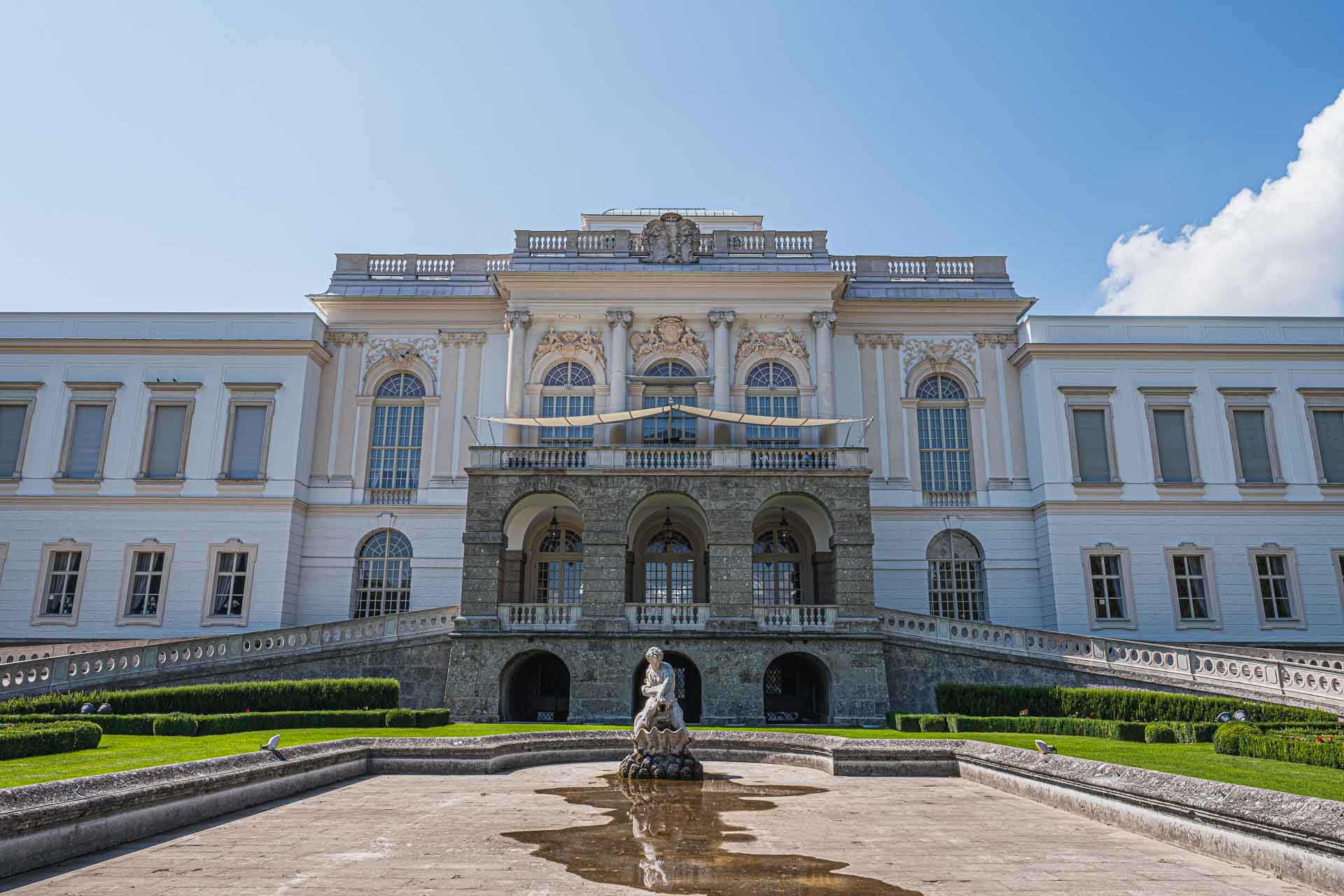 A grand neoclassical building in Salzburg with arched windows and a decorative facade, featuring a stone fountain with a statue in the foreground and manicured landscaping.