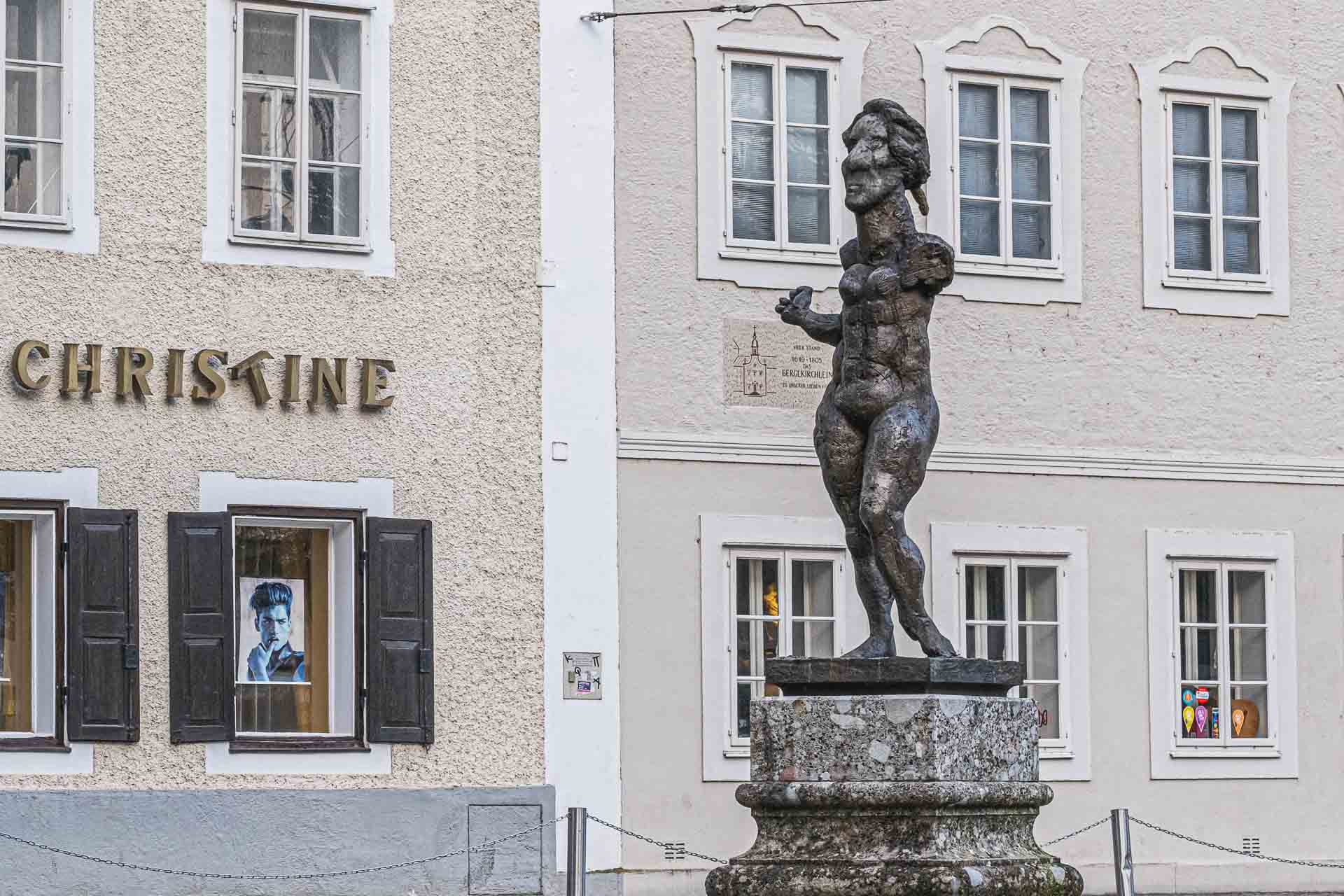 A statue of a muscular figure on a pedestal in front of an elegant building with the name "christine" and windows displaying various portraits.