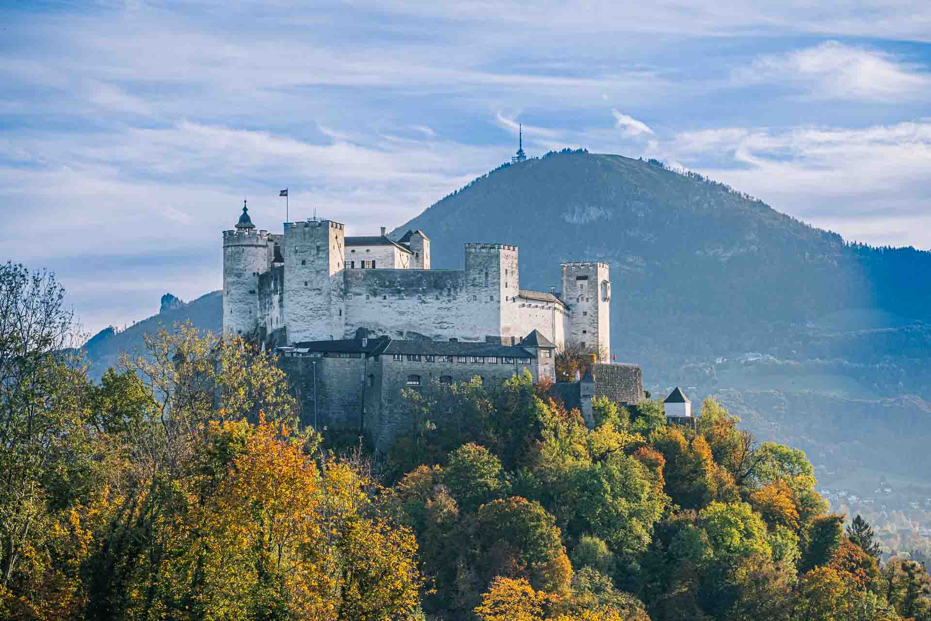 A medieval castle atop a hill surrounded by autumn foliage with mountains in the background.